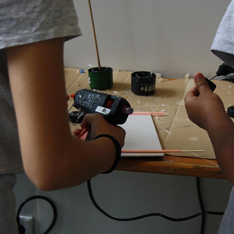 Zwei Kinder in grauen T‑Shirts an einem Tisch beim Basteln: links eine Hand hält eine Heißklebepistole, rechts eine Hand einen schwarzen Klebeaufsatz; auf dem Tisch Karton, Holzstäbchen, kleine Dosen und Klebereste.