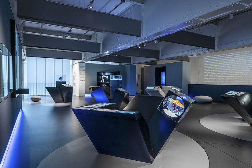 Im abgedunkelten Ausstellungsraum stehen angestrahlte Module zu jedem der zehn ausgestellten Projekte. Gestaltet mit kantigen Kuben.