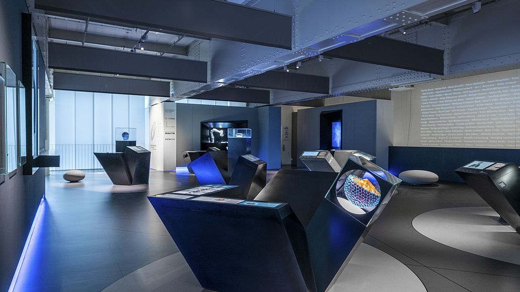Deutscher Zukunftspreis - Blick in die Ausstellung Im abgedunkelten Ausstellungsraum stehen angestrahlte Module zu jedem der zehn ausgestellten Projekte. Gestaltet mit kantigen Kuben.