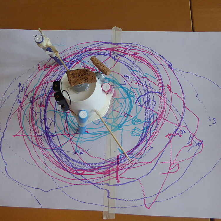 Selbstgebaute Kritzelmaschine mit Motor und Filzstiften auf Papier, die bunte, kreisförmige Linienmuster erzeugt.
