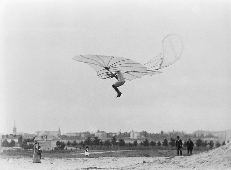 Otto Lilienthal von der Seite aufgenommen, die Beine angewinkelt, er fliegt im Segelapparat, unten auf einer Wiese stehen einige Zuschauer, im Hintergrund sieht man eine Stadt.