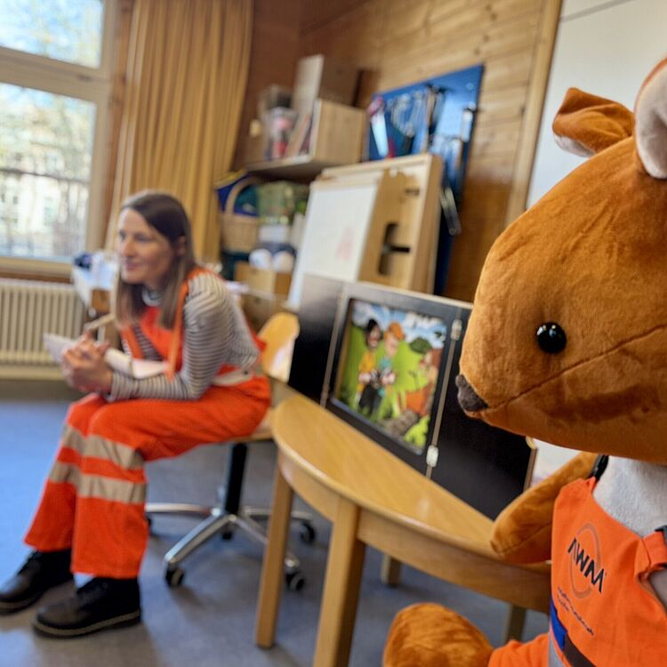 Links sitzt eine Müllwerkerin in orangener Weste, rechts ein großes, braunes Stoffeichhörnchen