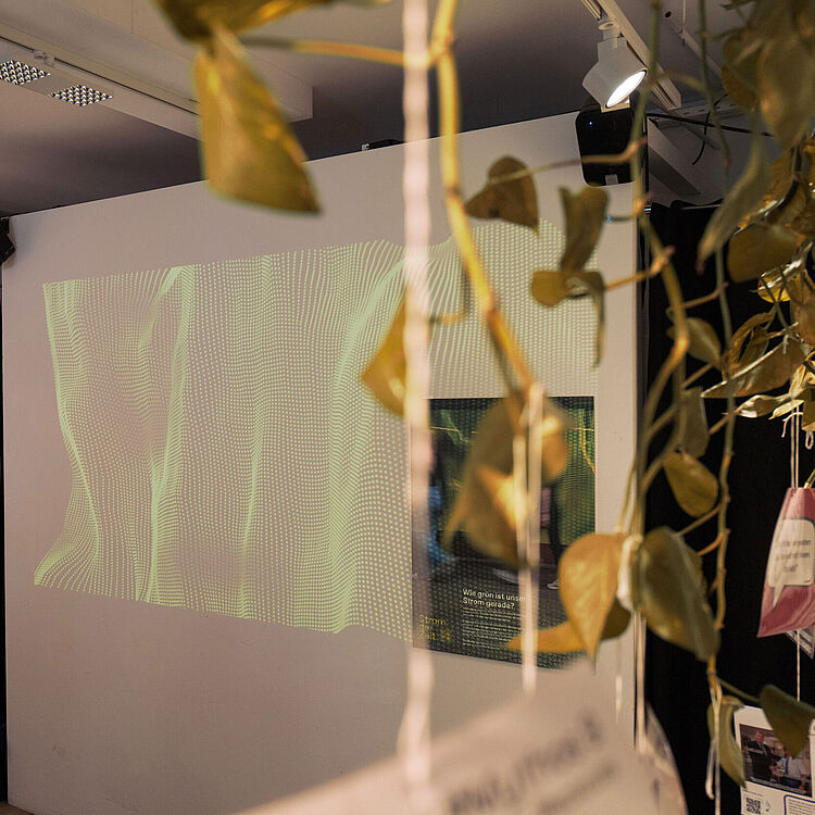 Ausstellungsraum mit einer Wandprojektion aus grünen Lichtmustern. Links hängt Kleidung unter einem Schild mit der Aufschrift ‚Was haben wir im Kleiderschrank?‘. Im Vordergrund sitzen zwei Personen, teilweise verdeckt sind Pflanzenblätter sichtbar.
