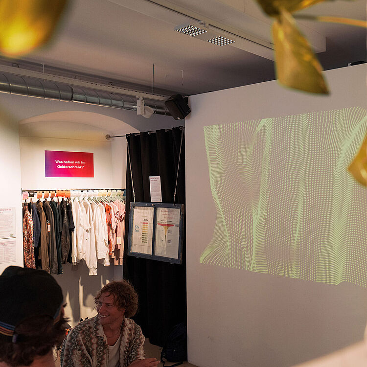 Ausstellungsraum mit einer Wandprojektion aus grünen Lichtmustern. Links hängt Kleidung unter einem Schild mit der Aufschrift ‚Was haben wir im Kleiderschrank?‘. Im Vordergrund sitzen zwei Personen, teilweise verdeckt sind Pflanzenblätter sichtbar.