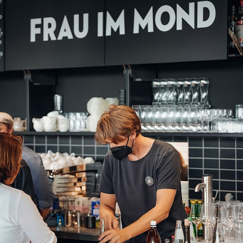 Selbstbedienungstheke im Restaurant mit einem Mitarbeiter und einer Besucherin. Im Hintergrund sieht man die Kaffeemaschine und viele Gläser.