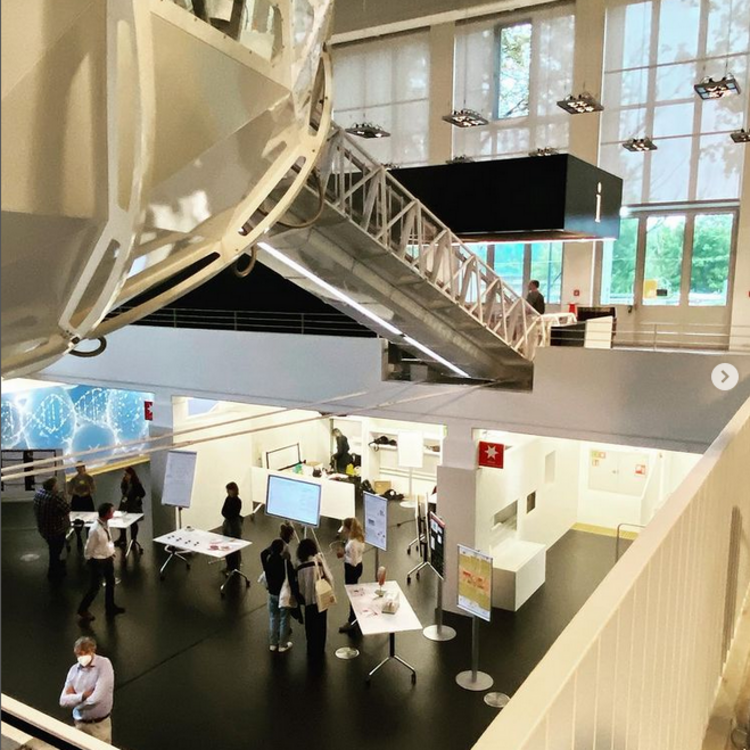 Innenaufnahme eines modernen, hellen Ausstellungsraums mit schwarzem Boden: im Vordergrund links oben ein großer weißer, kuppelartiger Struktorteil mit einer angelegten Metallbrücke/Steg, unten mehrere Stehtische, Poster-Displays und Touchscreens, Gruppen von Besuchern, Informationsständen und ein Mitarbeiterbereich im Hintergrund; hohe Fensterwände lassen Tageslicht herein.