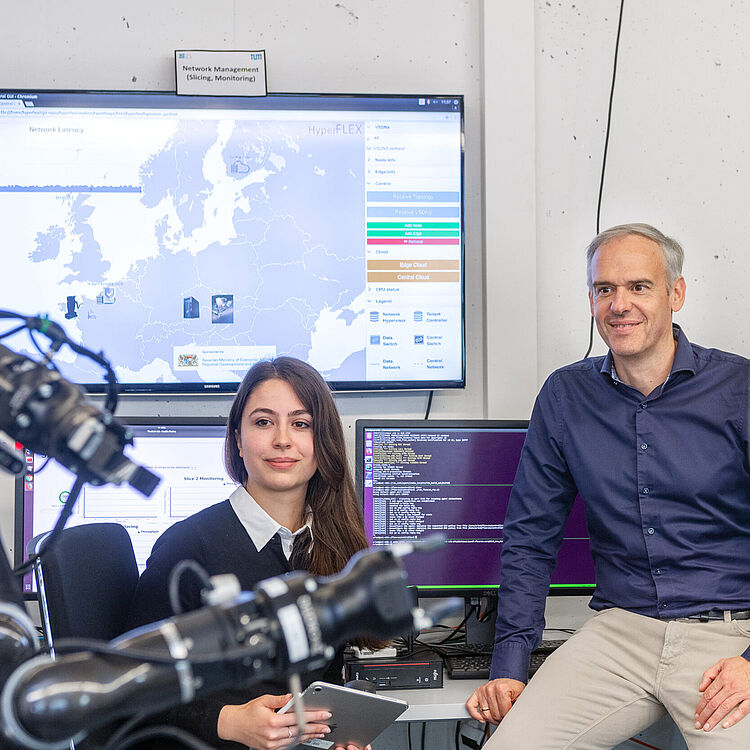 Zwei Personen in einem Laborbüro mit Monitoren und Roboterarmen: eine Frau sitzt mit Tablet, ein Mann lehnt an der Tischkante; im Hintergrund mehrere Bildschirme zeigen eine Europakarte mit Netzwerkdaten, Terminalausgaben und ein Schild „Network Management (Slicing, Monitoring)“.