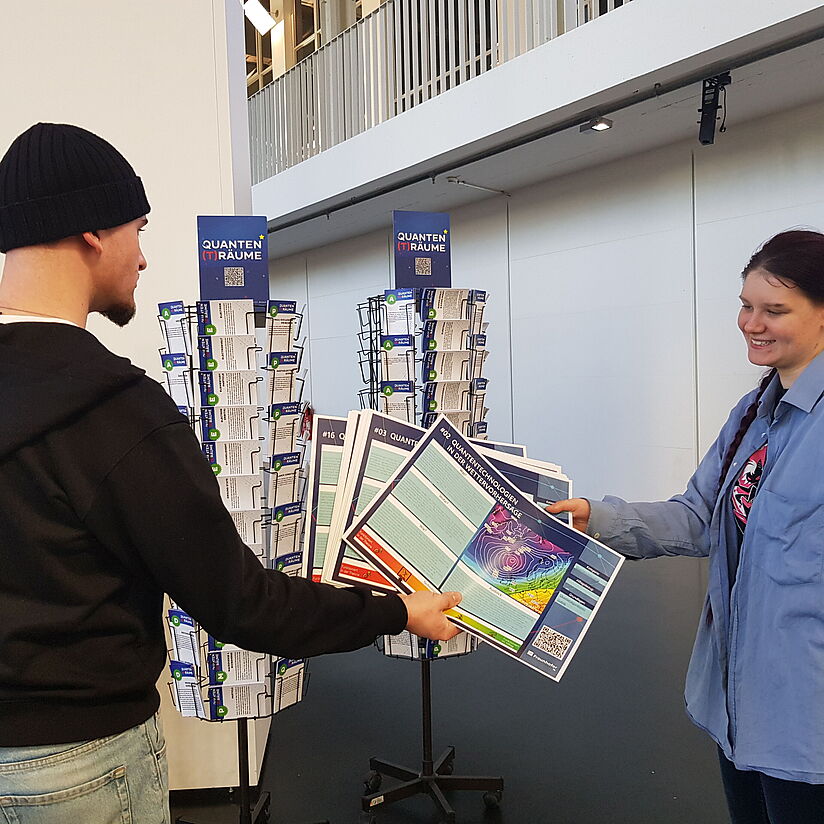 Informationskarten werden von einer Person an eine andere übergeben. Im Hintergrund sind weitere Materialien auf einem Postkatenständer.
