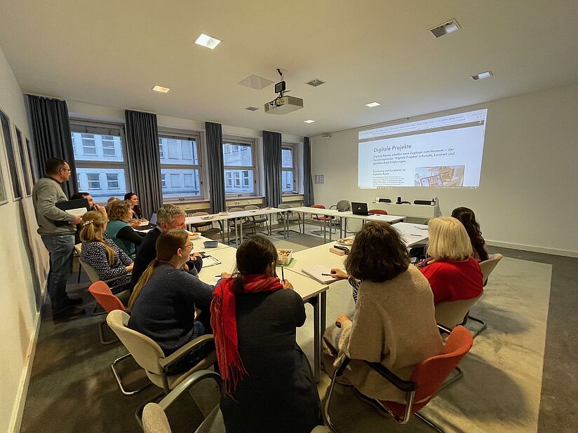 In einem Seminarraum sitzen Frauen und Männer an Besprechungstischen und folgen einer Präsentation, die an eine Wand projiziert wird.