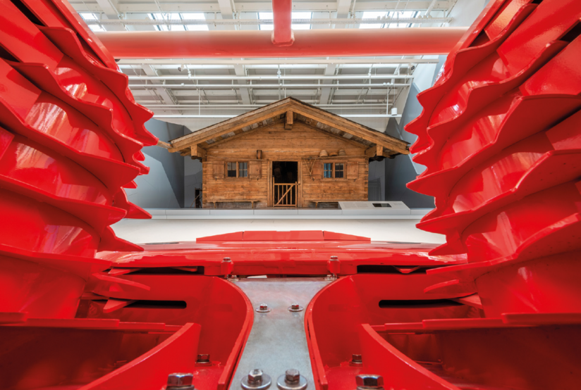 Ein Gerät aus leuchtend rotem Metall durch das hindurch eine hölzerne Hütte zu sehen ist.