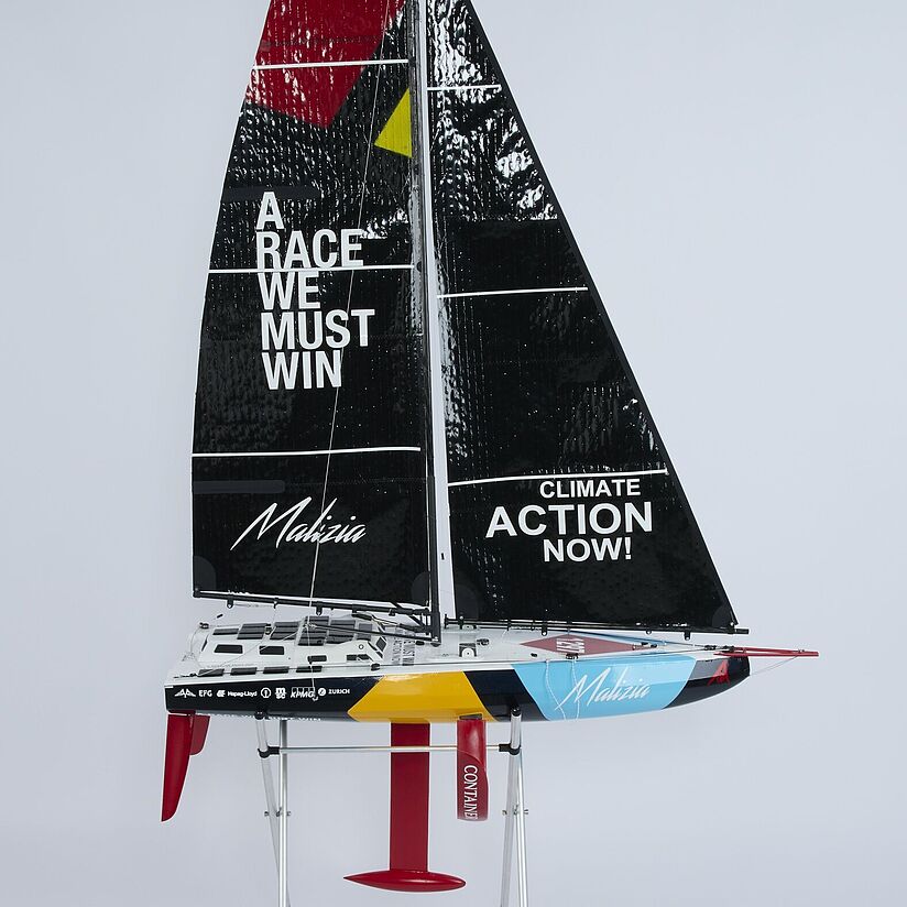 Modell eines Rennseglers auf Ständer mit schwarzem Großsegel, Schriftzug „A RACE WE MUST WIN“ links und „CLIMATE ACTION NOW!“ rechts, bunte Rumpfbemalung und roter Kiel.