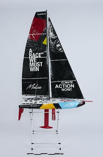 Modell eines Rennseglers auf Ständer mit schwarzem Großsegel, Schriftzug „A RACE WE MUST WIN“ links und „CLIMATE ACTION NOW!“ rechts, bunte Rumpfbemalung und roter Kiel.