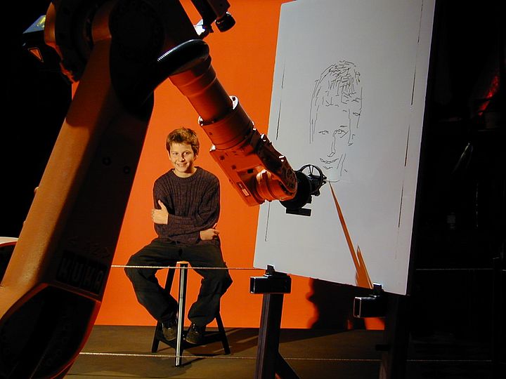 Junge sitzt Modell für einen Roboter. Ein Roboterarm zeichnet ein Portrait des Jungen auf eine Leinwand.