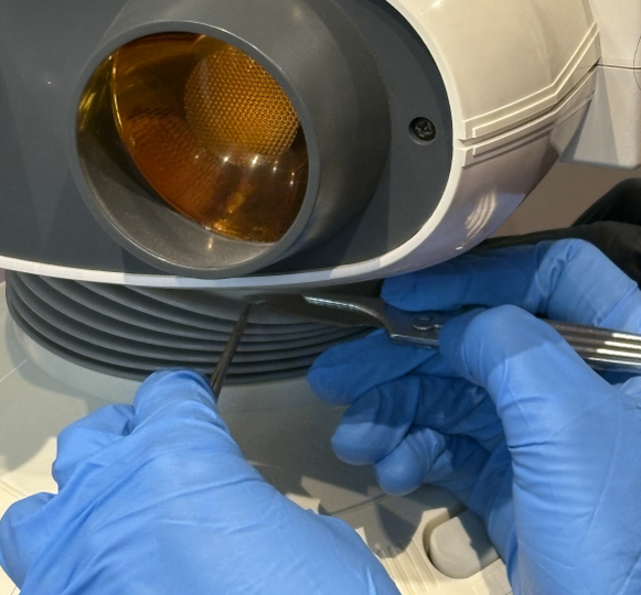 Zwei Hände in blauen Handschuhen mit Pinzette vor optischem Instrument