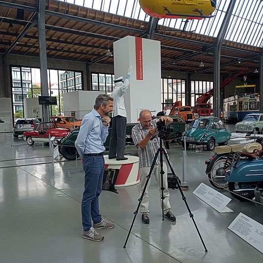 Fotoshooting im Verkehrszentrum: Zwei Männer betrachten Aufnahmen im Display einer Fotokamera umgeben von Oltimern