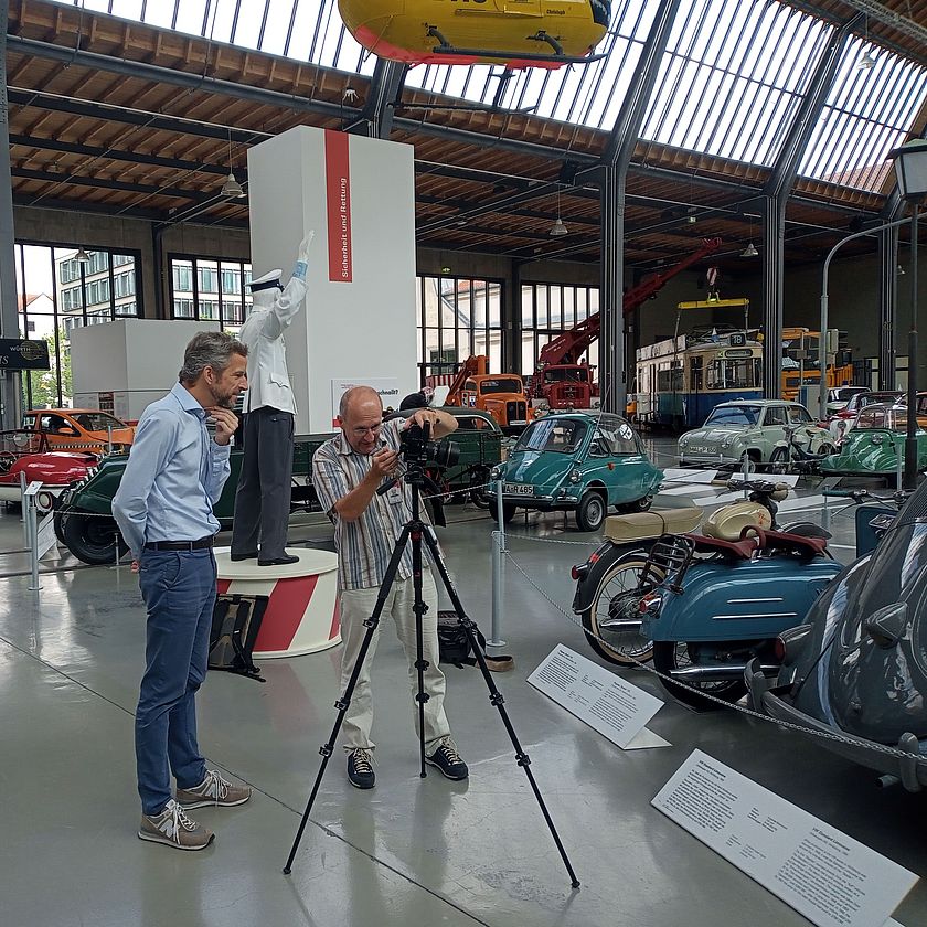 Fotoshooting im Verkehrszentrum: Zwei Männer betrachten Aufnahmen im Display einer Fotokamera umgeben von Oltimern