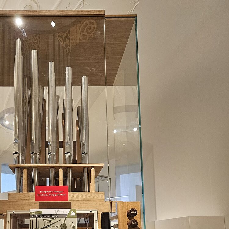 Vitrine mit historischem Orgelprospekt und sichtbaren Metallpfeifen auf Holzgestell, darunter Infotafel und fünf runde Registerknöpfe, im hellen Museumssaal.