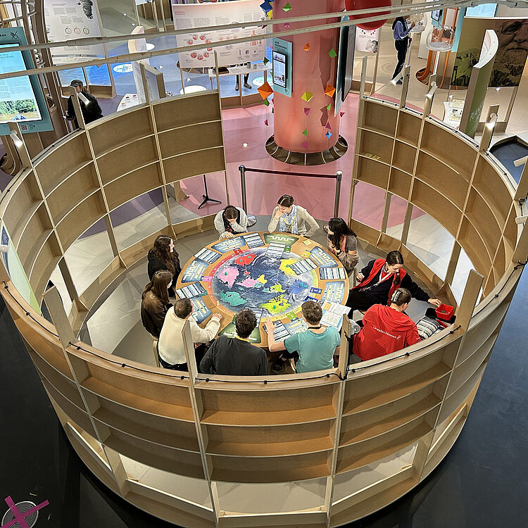 Zehn Menschen sitzen an einem runden Tisch, auf dem eine große Weltkarte abgebildet ist und spielen ein Spiel.