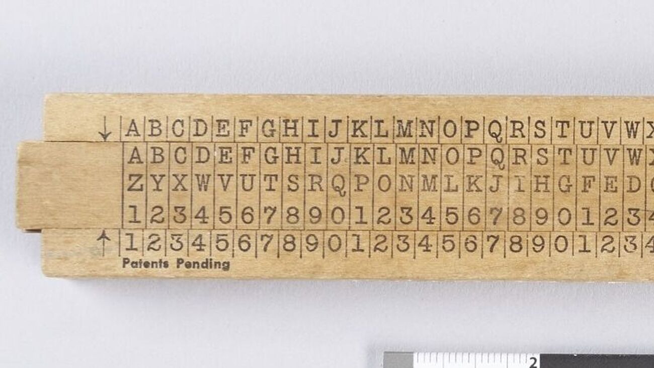 Holzschieber. Fest oben Alphabet zweifach, unten wiederholte Zahlenfolge 0 bis 9. Mittig verschiebbar drei Zeilen: Alphabet, Alphabet rückläufig, Zahlenfolge 0 bis 9