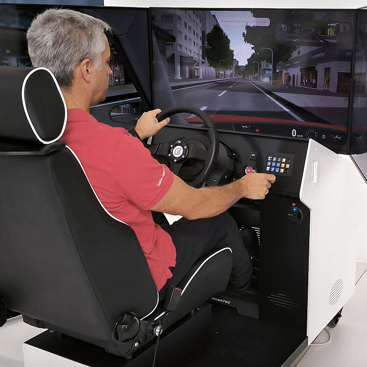 Ein Mann sitzt in einem modernen Fahrsimulator mit einem großen gebogenen Bildschirm. Er hält das Lenkrad und bedient ein Tastenfeld. Auf dem Bildschirm ist eine virtuelle Stadtstraße zu sehen.