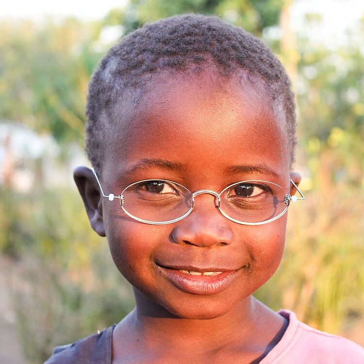 Nahaufnahme eines Kindes mit kurzem, lockigem Haar, rosa T‑Shirt und silberner Brille, freier Naturhintergrund mit unscharfen Bäumen und Büschen.