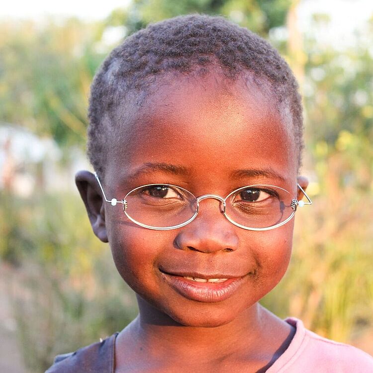 Nahaufnahme eines Kindes mit kurzem, lockigem Haar, rosa T‑Shirt und silberner Brille, freier Naturhintergrund mit unscharfen Bäumen und Büschen.