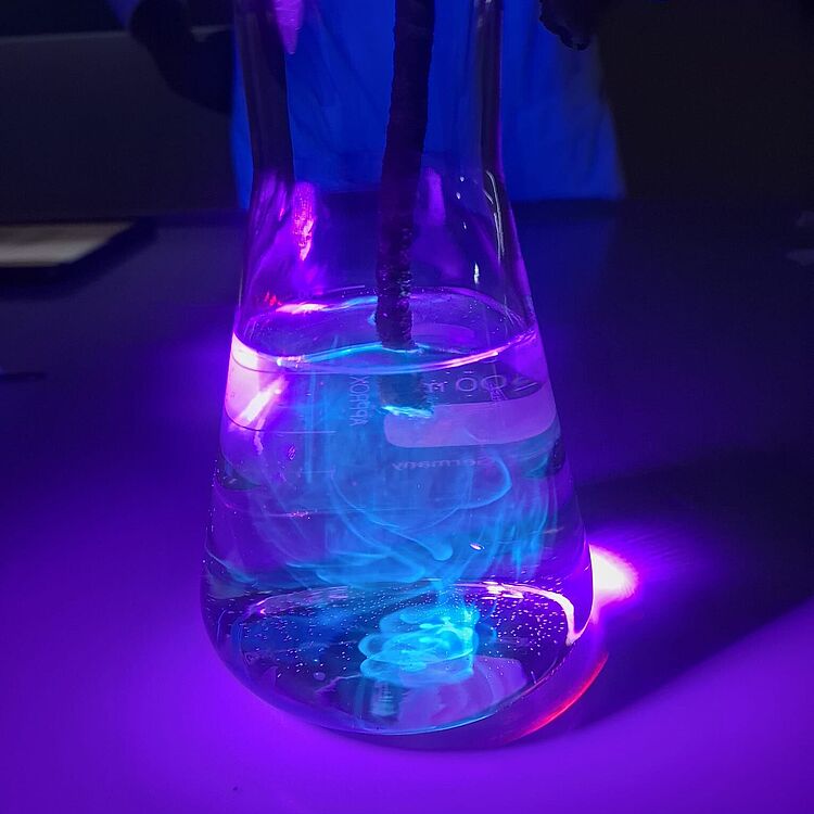 Erlenmeyerkolben auf weißer Oberfläche, gefüllt mit klarem Wasser und einem dunklen Stab, blau-türkisfarbene Wirbel und Nebel im Inneren, unter violettem UV-/Neonlicht leuchtend.