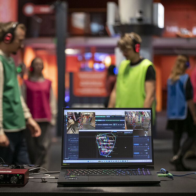 Ein Laptop auf einem Tisch zeigt eine Gesichts-Tracking-Software mit markierten Punkten; im unscharfen Hintergrund mehrere Personen in bunten Sicherheitswesten und kabellosen Kopfhörern bewegen sich in einem orange beleuchteten, musealen oder Event-Raum.