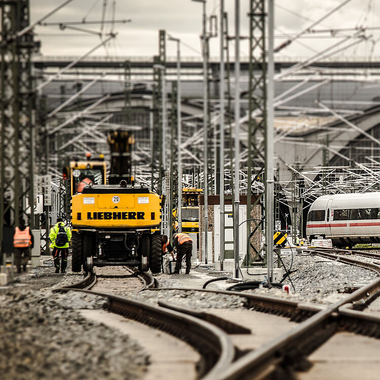 Blick auf eine Bahnbaustelle. Gelbe Bagger und Bauarbeiter arbeiten an den Gleisen, während im Hintergrund ein ICE-Zug vorbeifährt.