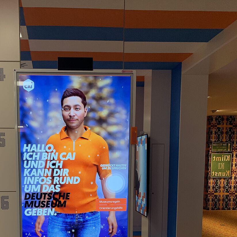 Interaktives Digitaldisplay mit einer anonymisierten männlichen Figur in orange Polohemd und Jeans, umgeben von blau-orange gestreiftem Interieur; linker Bildrand zeigt nummerierte Schließfächer (134–137), Boden Kachelmuster in Blau, Orange und Beige; Text auf Bildschirm: „Hallo, ich bin CAI und ich kann dir Infos rund um das Deutsche Museum geben.“