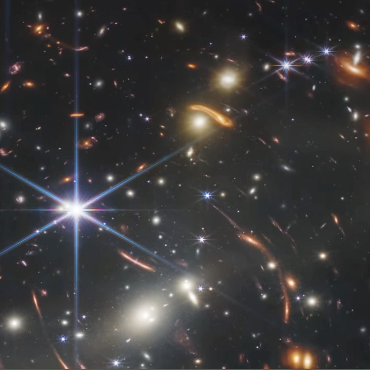 Das James-Webb-Weltraumteleskop-Feld mit tausenden fernen Galaxien und sternartigen Spikes: helles, achtstrahliges Sternsignal in Bildmitte, zahlreiche vergrößert verzerrte orangefarbene Galaxienbögen (Gravitationslinsen), blaue und weiße Punktquellen und diffuse Galaxien vor einem schwarzen Weltraumhintergrund.