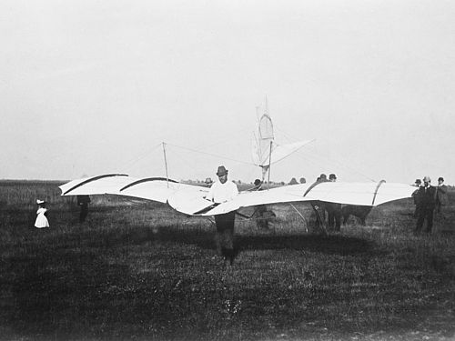 Otto Lilienthal steht auf einer Wiese und trägt den Gleiter fertig aufgebaut.t