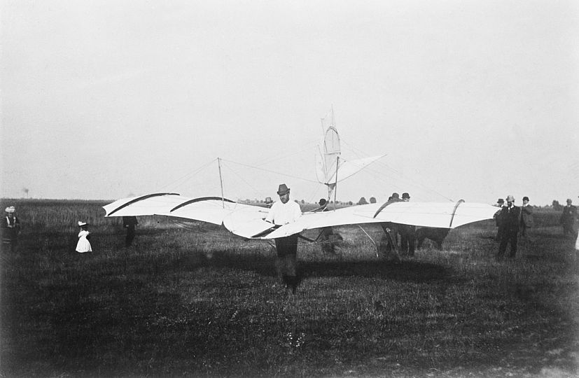 Otto Lilienthal steht auf einer Wiese und trägt den Gleiter fertig aufgebaut.t 